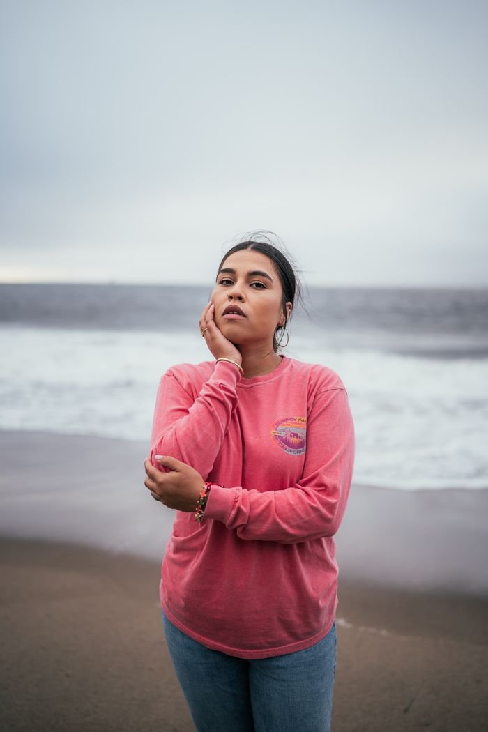 Woman in a pink sweater posing calmly by the beach with a muted, overcast sky.
