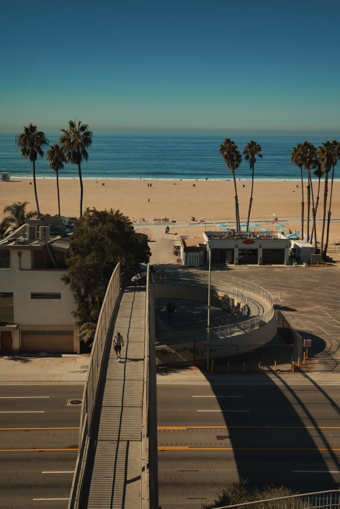 Explore the sunlit Santa Monica beach and urban footbridge from above, capturing coastal beauty.