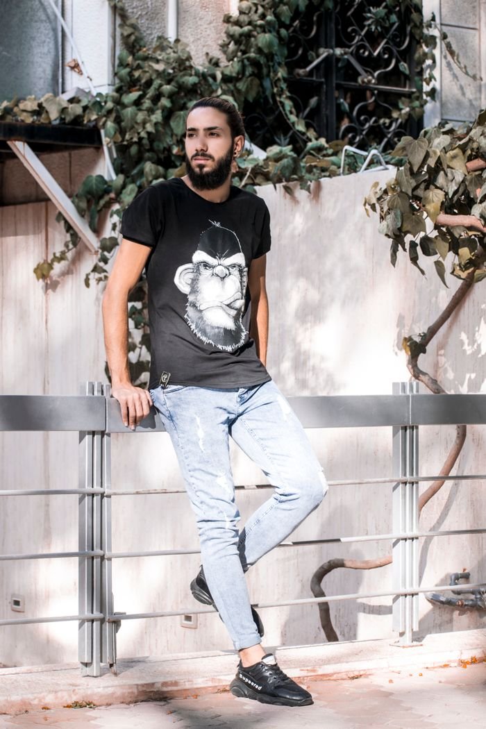 A fashionable man in a casual outfit leaning on a fence outside.