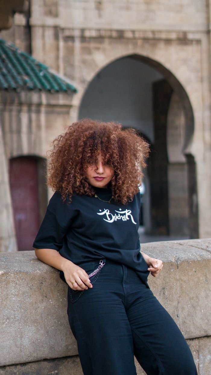 Stylish young woman poses against Moroccan architectural backdrop, showcasing curly hair and fashion sense.