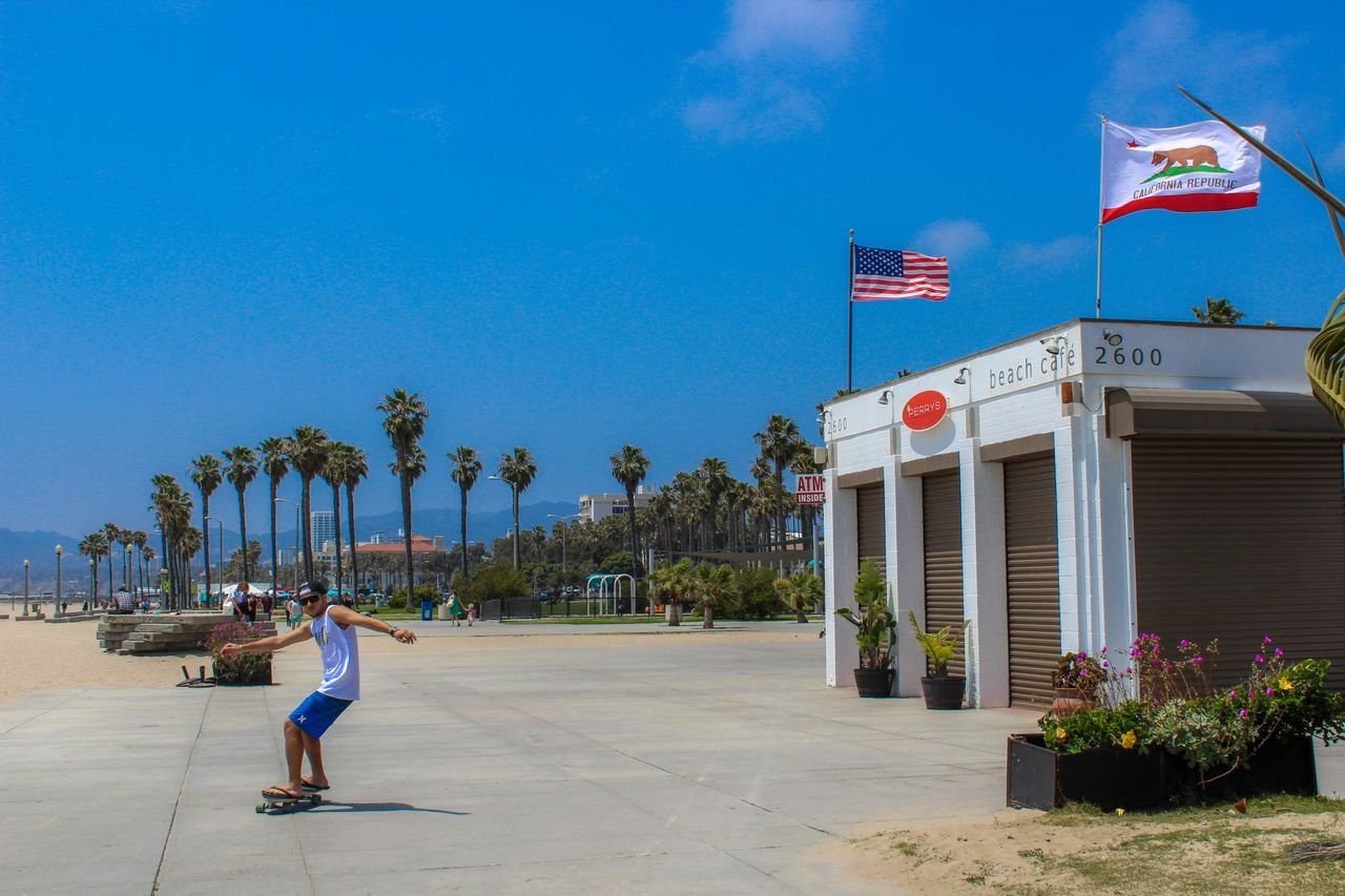 A skateboarder glides past a beach cafe with palm trees under a clear blue sky.